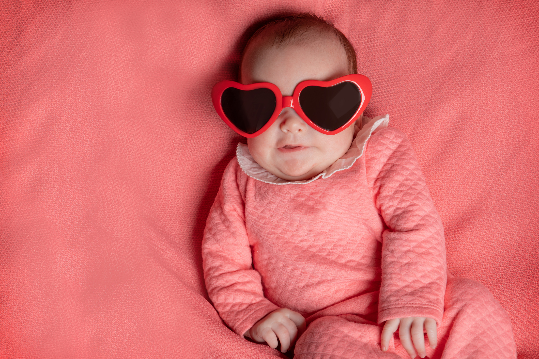 A portrait of an adorable 3 month old baby girl chillin' on a pink blanket while wearing her heart shaped glasses for Valentine's Day.